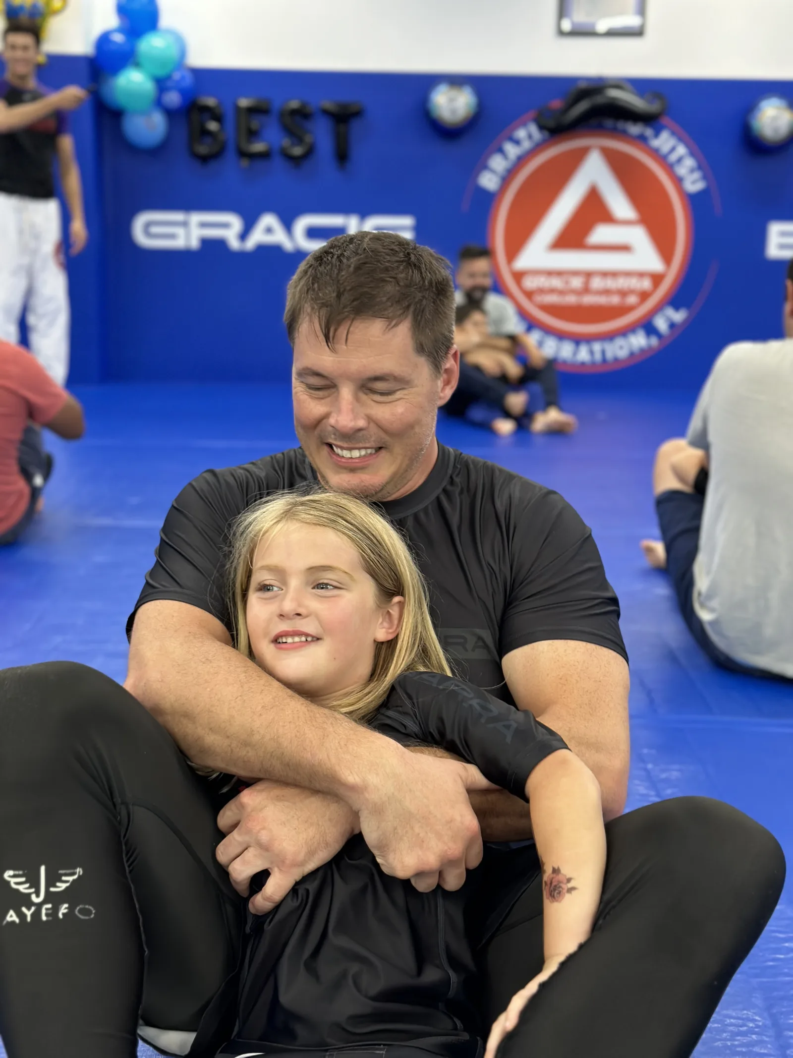 Dad sitting on the mat at Gracie Barra Celebration holding his young daughter during a parents-and-kids class